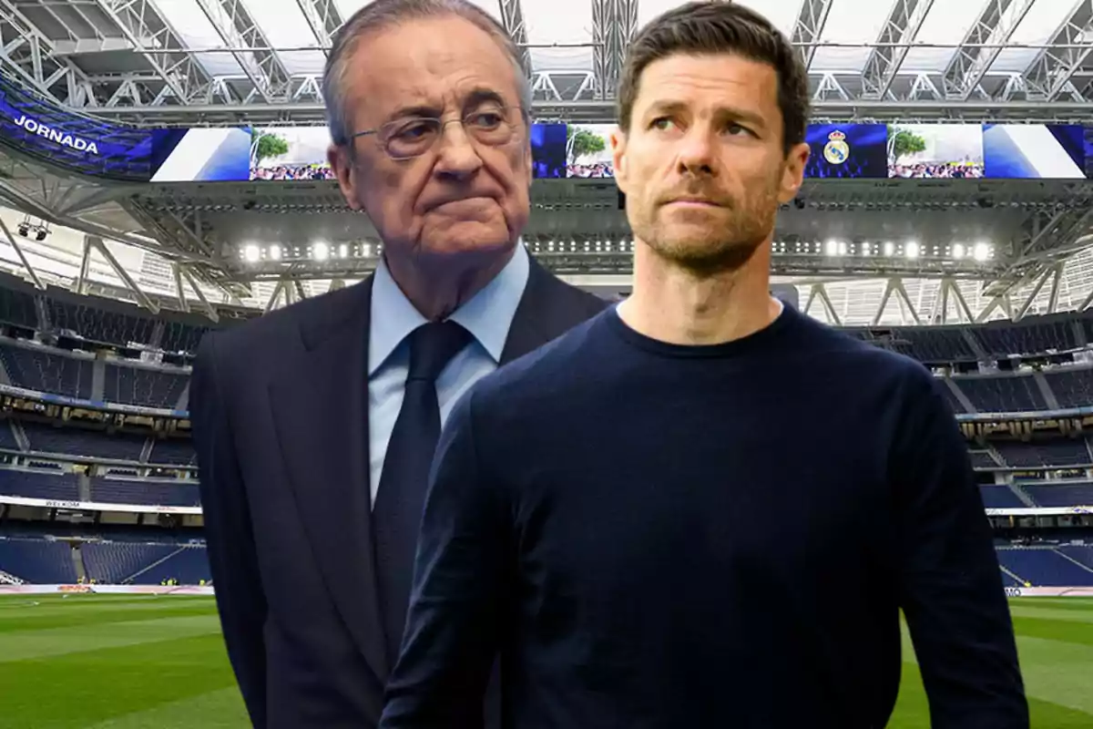 Dos hombres de pie en el interior de un estadio de fútbol moderno, uno con traje y gafas y el otro con suéter oscuro, ambos mirando en direcciones opuestas.