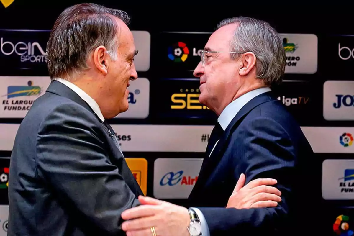 Dos hombres de traje se saludan cordialmente frente a un fondo con logotipos de patrocinadores deportivos