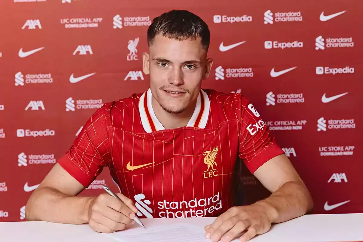 Soccer player wearing a Liverpool jersey signing a contract in front of a red background with sponsor logos. Soccer player wearing a Liverpool jersey signing a contract in front of a red background with sponsor logos.