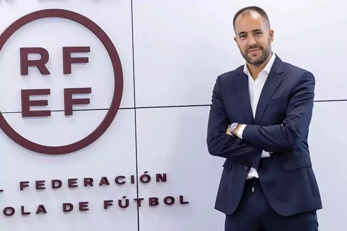 Man in a blue suit posing with his arms crossed next to a Royal Spanish Football Federation logo on a white wall Man in a blue suit posing with his arms crossed next to a Royal Spanish Football Federation logo on a white wall