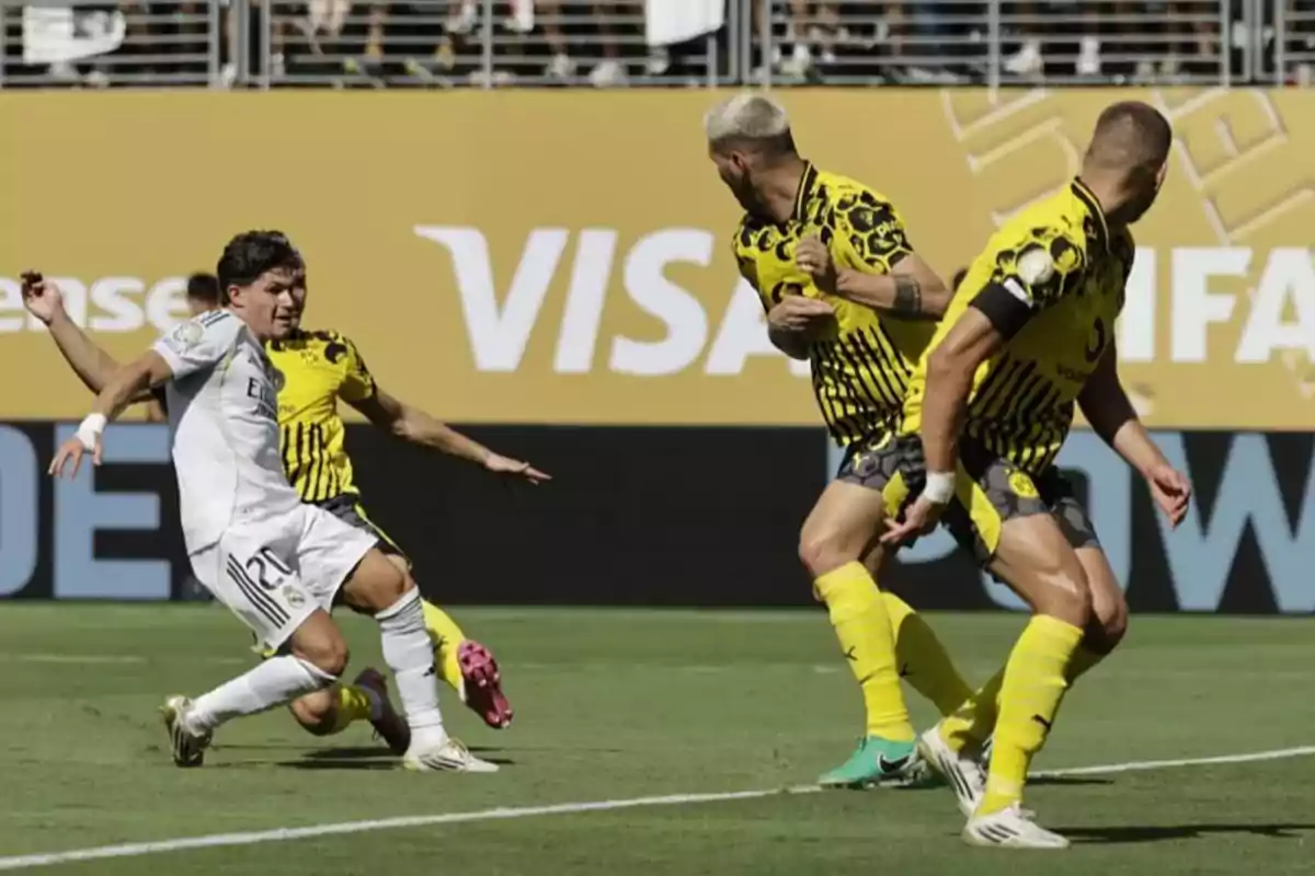 Un jugador de fútbol con uniforme blanco intenta disparar el balón mientras dos defensores con uniforme amarillo y negro intentan bloquearlo durante un partido en un estadio. Un jugador de fútbol con uniforme blanco intenta disparar el balón mientras dos defensores con uniforme amarillo y negro intentan bloquearlo durante un partido en un estadio.