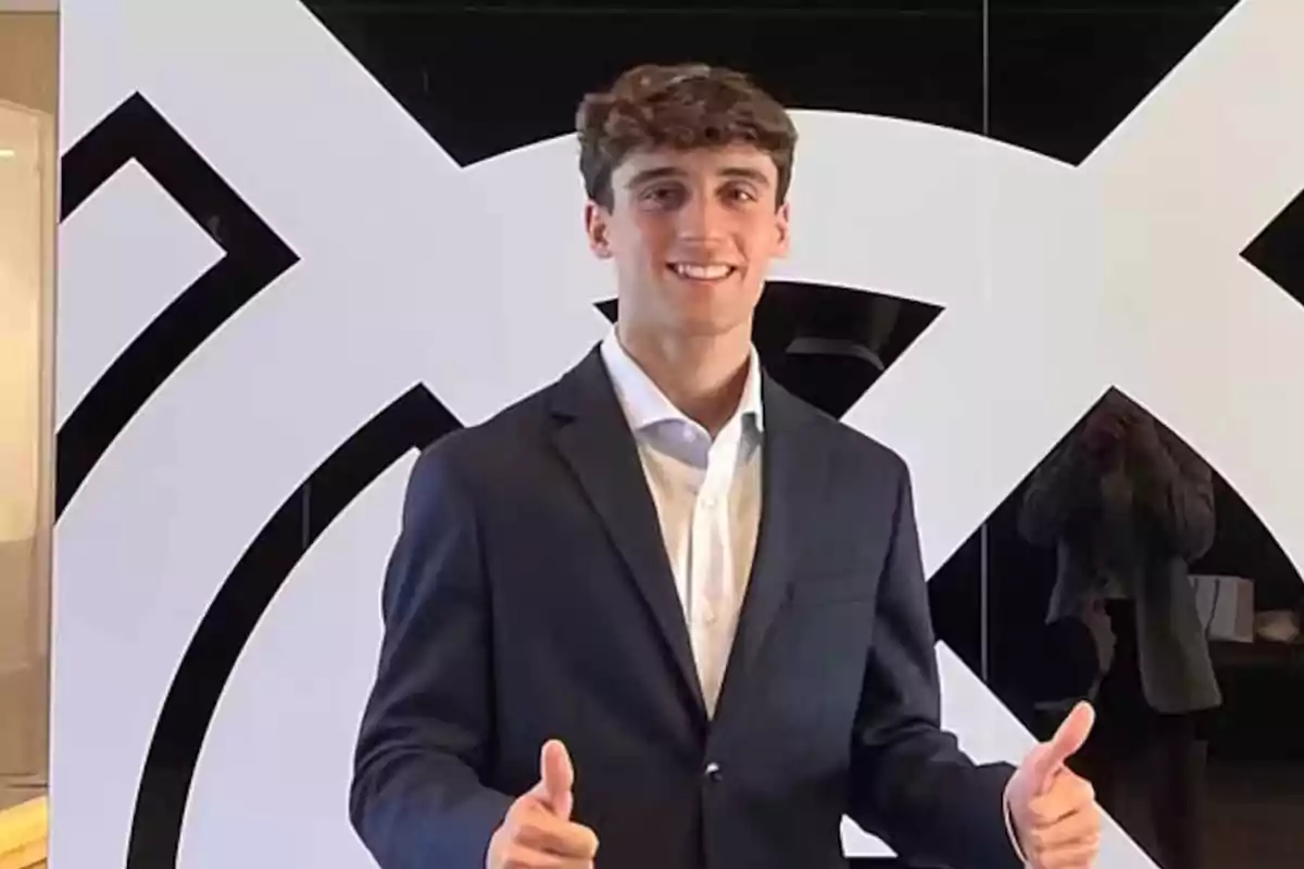 Joven con traje oscuro y camisa blanca sonriendo y levantando ambos pulgares frente a un fondo con diseño geométrico en blanco y negro Joven con traje oscuro y camisa blanca sonriendo y levantando ambos pulgares frente a un fondo con diseño geométrico en blanco y negro