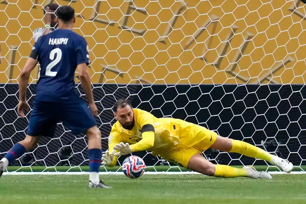 Portero vestido de amarillo atajando un balón durante un partido de fútbol mientras un defensor observa la jugada dentro del área