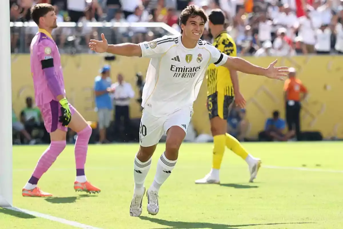 Futbolista del Real Madrid celebrando un gol con los brazos abiertos mientras un portero y otro jugador observan en el fondo durante un partido diurno Futbolista del Real Madrid celebrando un gol con los brazos abiertos mientras un portero y otro jugador observan en el fondo durante un partido diurno