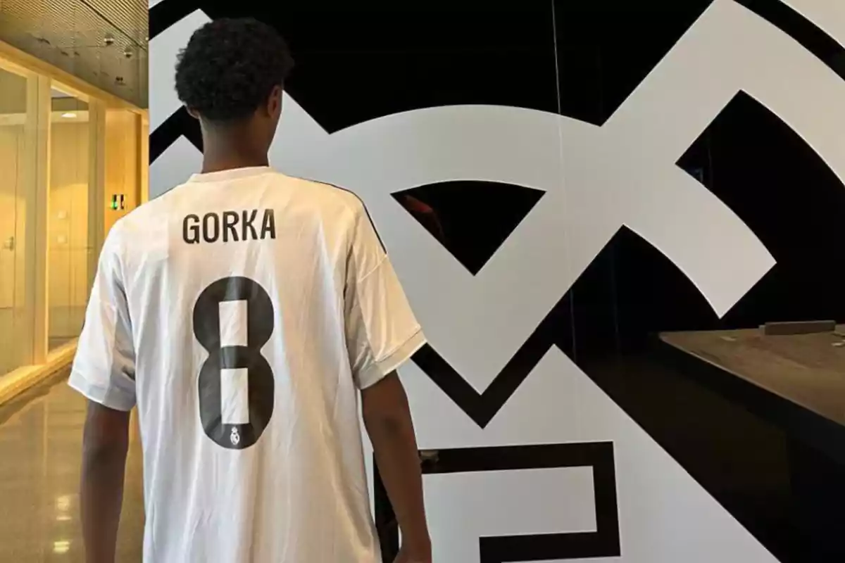 Persona de espaldas con camiseta blanca del Real Madrid con el nombre Gorka y el número 8 frente a un gran logo del club Persona de espaldas con camiseta blanca del Real Madrid con el nombre Gorka y el número 8 frente a un gran logo del club