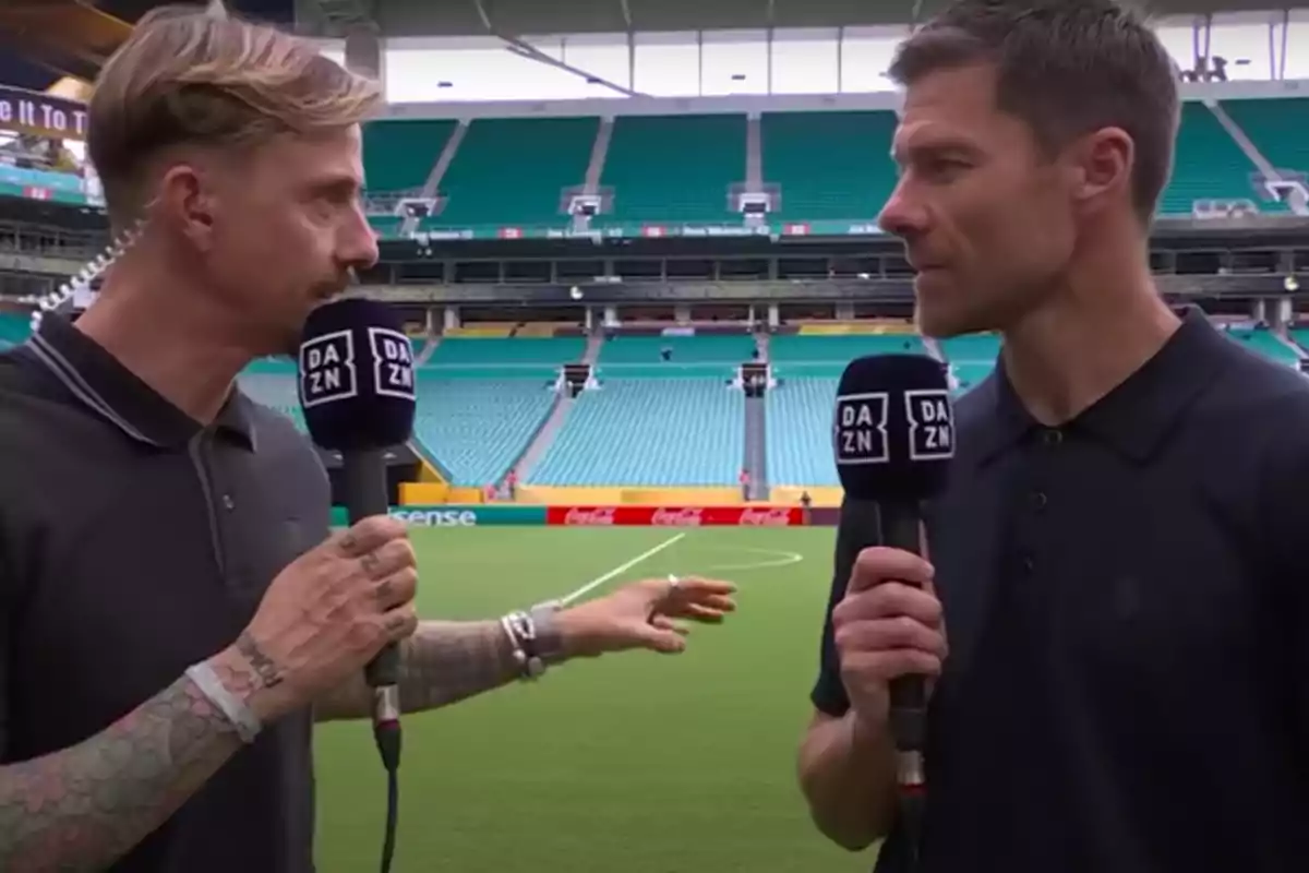 Dos hombres con micrófonos de DAZN conversan en el campo de un estadio de fútbol vacío