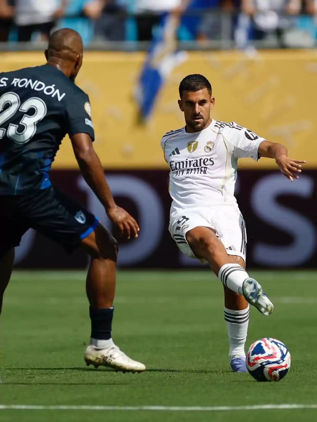Dos futbolistas disputan el balón durante un partido en un estadio, uno con uniforme blanco y el otro con uniforme azul oscuro Dos futbolistas disputan el balón durante un partido en un estadio, uno con uniforme blanco y el otro con uniforme azul oscuro