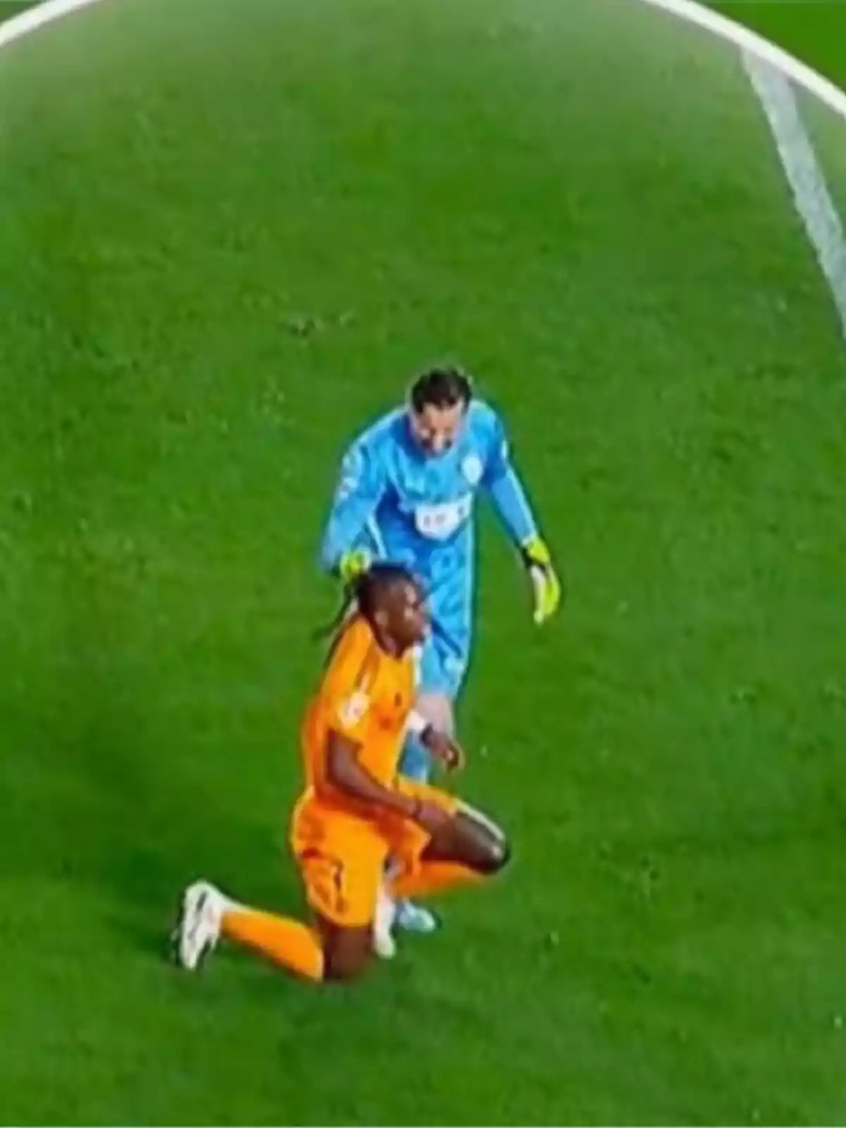 Un portero con uniforme azul y guantes amarillos está detrás de un jugador de fútbol con uniforme naranja que está arrodillado en el césped durante un partido Un portero con uniforme azul y guantes amarillos está detrás de un jugador de fútbol con uniforme naranja que está arrodillado en el césped durante un partido