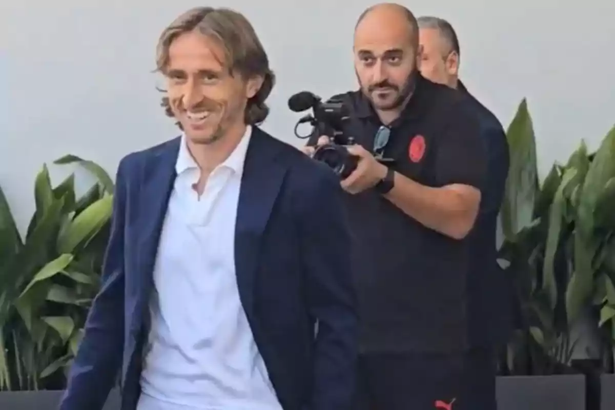 Hombre sonriente con chaqueta azul y camisa blanca caminando mientras es grabado por un camarógrafo, con plantas verdes al fondo. Hombre sonriente con chaqueta azul y camisa blanca caminando mientras es grabado por un camarógrafo, con plantas verdes al fondo.