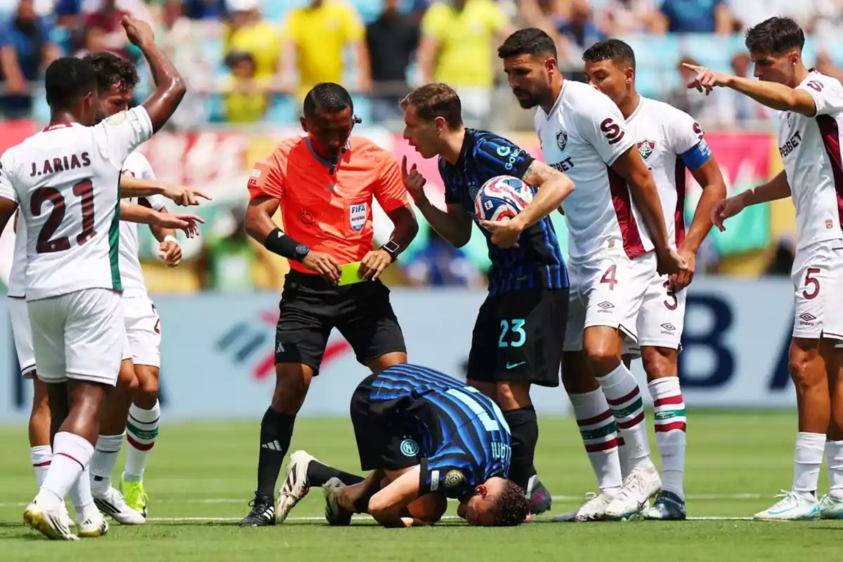Un árbitro muestra una tarjeta amarilla mientras varios jugadores discuten y uno de ellos está en el suelo durante un partido de fútbol Un árbitro muestra una tarjeta amarilla mientras varios jugadores discuten y uno de ellos está en el suelo durante un partido de fútbol