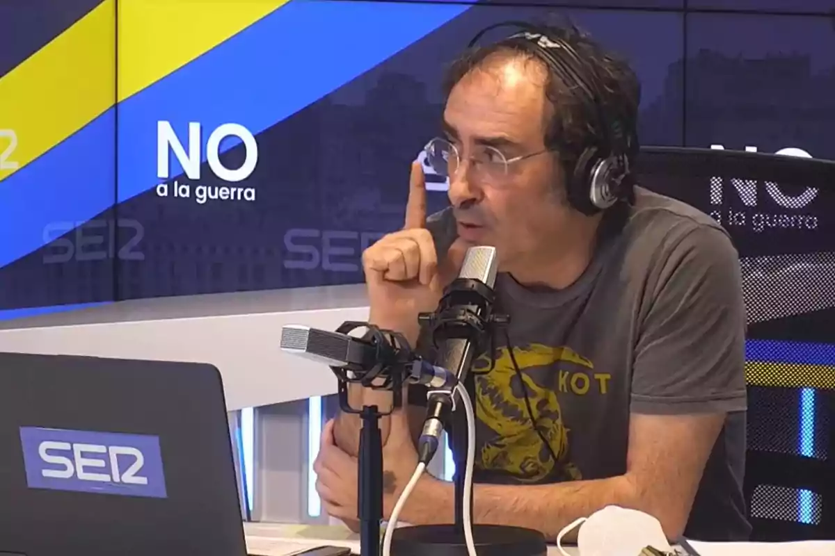Hombre con auriculares y camiseta gris hablando frente a un micrófono en un estudio de radio con un cartel de fondo que dice NO a la guerra