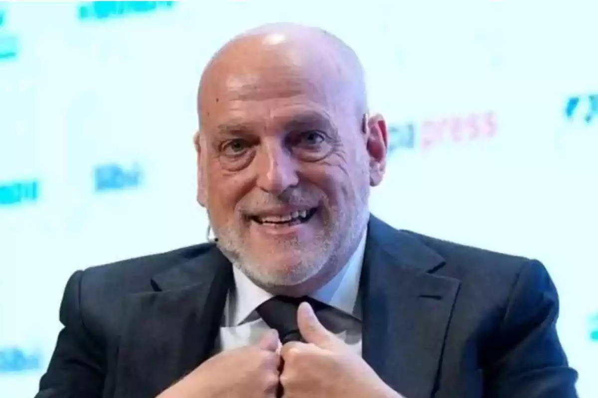 Hombre calvo de mediana edad con barba canosa sonriendo y usando traje oscuro mientras ajusta su corbata en un entorno profesional.