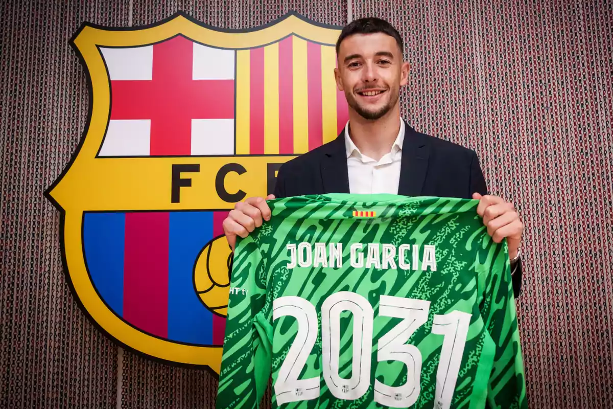 Un hombre sonriente sostiene una camiseta de portero verde con el nombre Joan Garcia y el número 2031 frente al escudo del FC Barcelona. Un hombre sonriente sostiene una camiseta de portero verde con el nombre Joan Garcia y el número 2031 frente al escudo del FC Barcelona.