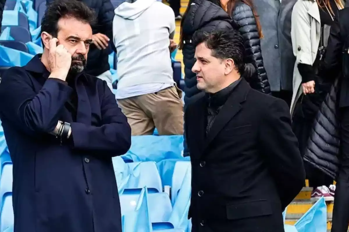 Two men are standing and talking in the stands of a stadium; one of them is holding his hand near his face while the other watches him. Both are wearing dark coats, and in the background, more people and blue seats can be seen. Two men are standing and talking in the stands of a stadium; one of them is holding his hand near his face while the other watches him. Both are wearing dark coats, and in the background, more people and blue seats can be seen.