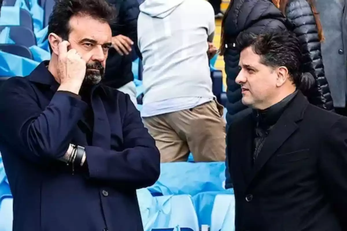 Dos hombres conversan en las gradas de un estadio, ambos vestidos con ropa oscura y rodeados de personas con abrigos. Dos hombres conversan en las gradas de un estadio, ambos vestidos con ropa oscura y rodeados de personas con abrigos.