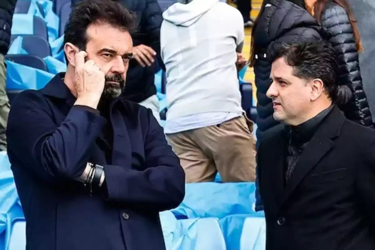 Dos hombres de pie conversan en un estadio con asientos azules mientras uno de ellos sostiene su mano cerca de la cara Dos hombres de pie conversan en un estadio con asientos azules mientras uno de ellos sostiene su mano cerca de la cara