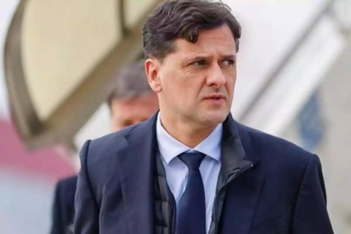 Hombre de cabello corto y oscuro con traje azul y corbata, mirando hacia un lado en un entorno exterior Hombre de cabello corto y oscuro con traje azul y corbata, mirando hacia un lado en un entorno exterior