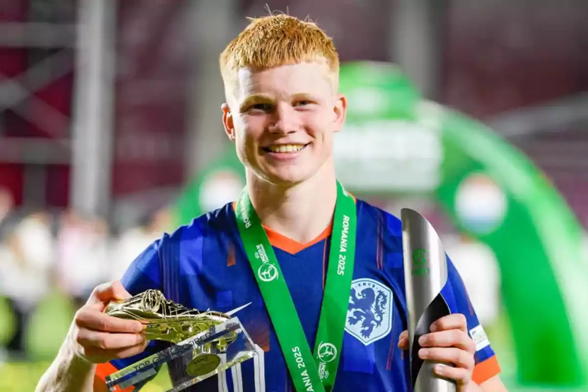 Joven futbolista sonriente con uniforme azul sostiene un trofeo dorado y una medalla verde en un estadio Joven futbolista sonriente con uniforme azul sostiene un trofeo dorado y una medalla verde en un estadio