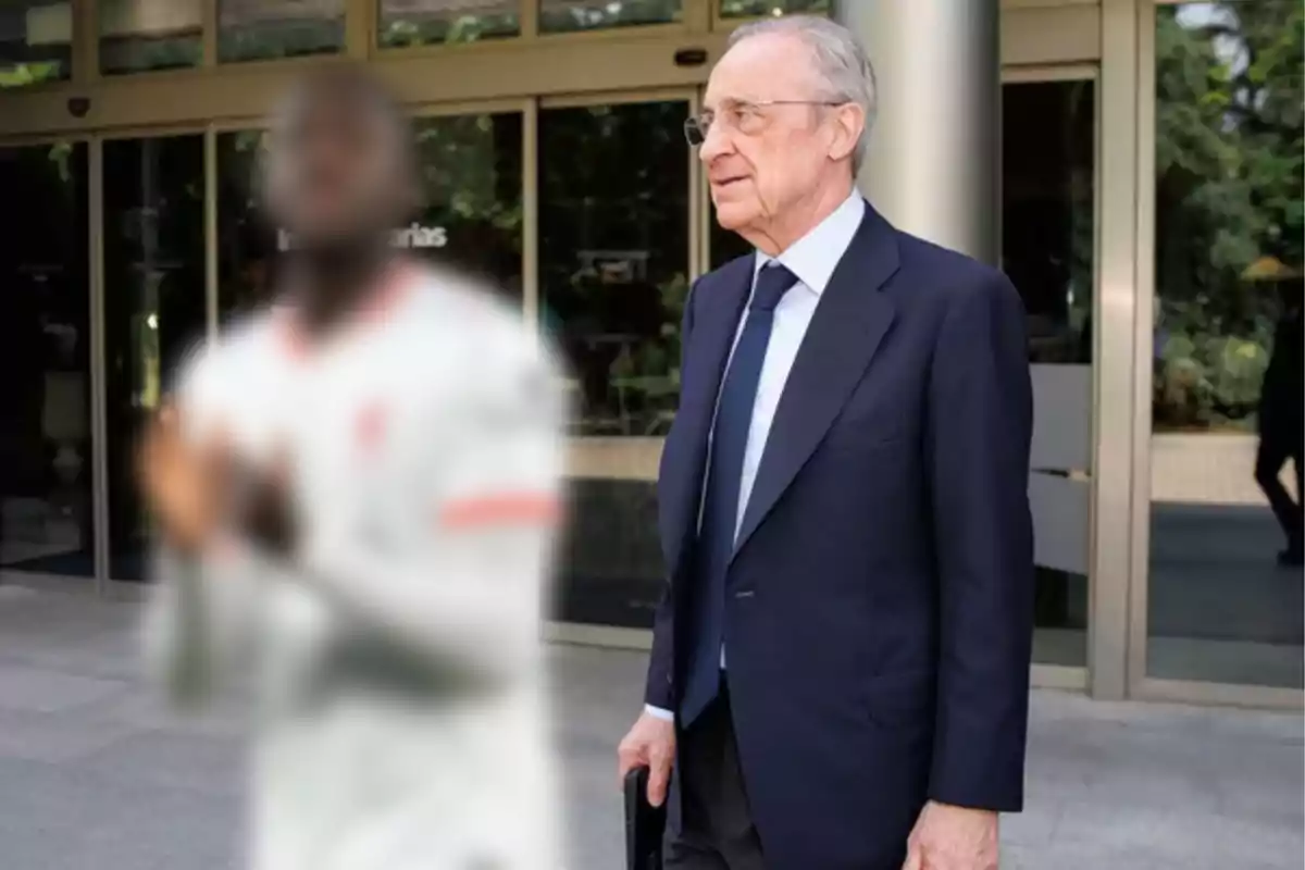 Man in a blue suit standing in front of a building next to a blurred person in a white jersey