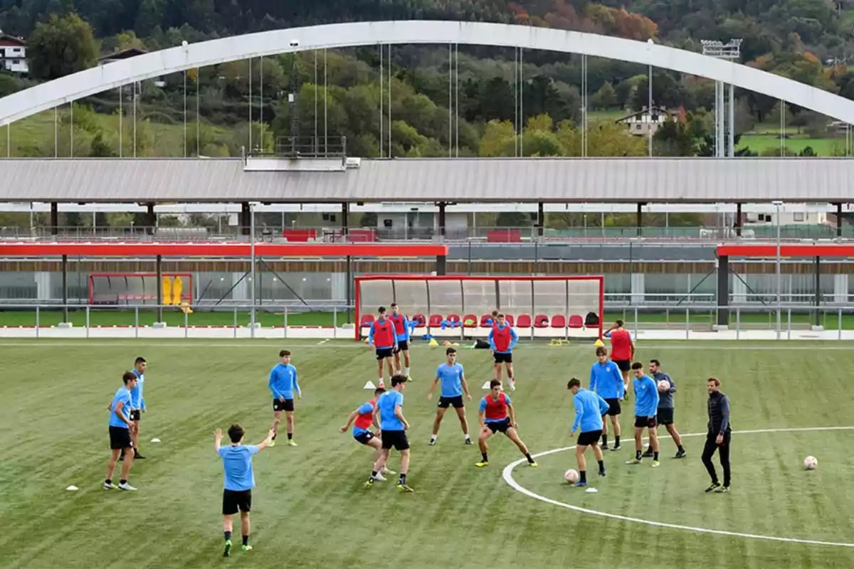 Jugadores de fútbol entrenando en un campo de césped con entrenadores observando y una estructura de estadio al fondo Jugadores de fútbol entrenando en un campo de césped con entrenadores observando y una estructura de estadio al fondo