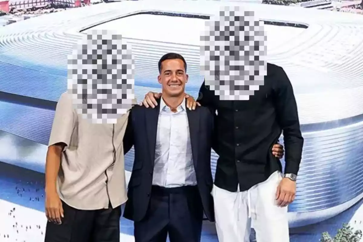 Three people pose in front of a stadium model, two of them with pixelated faces, and the person in the center smiles wearing a suit.