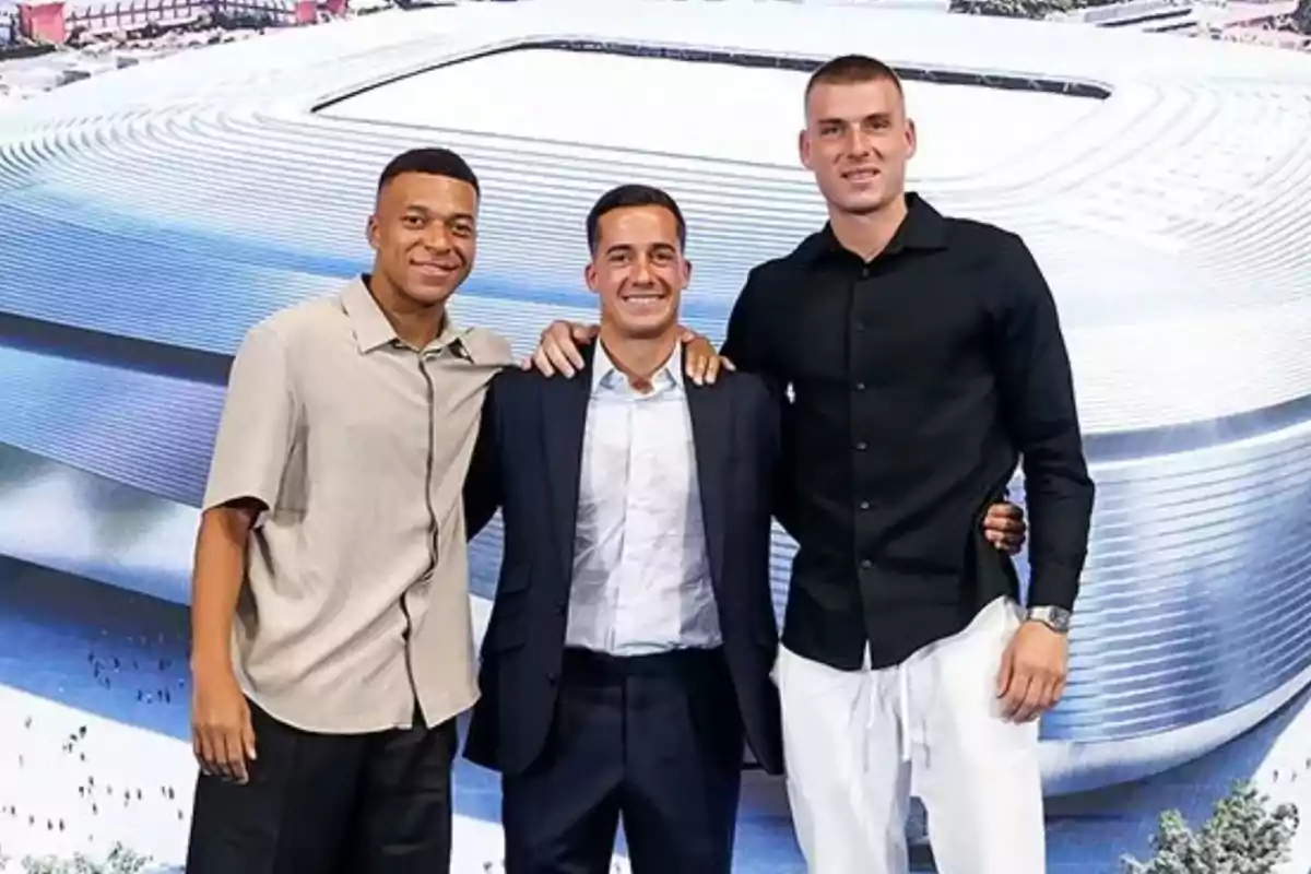 Tres hombres posan juntos sonriendo frente a una imagen de un estadio moderno