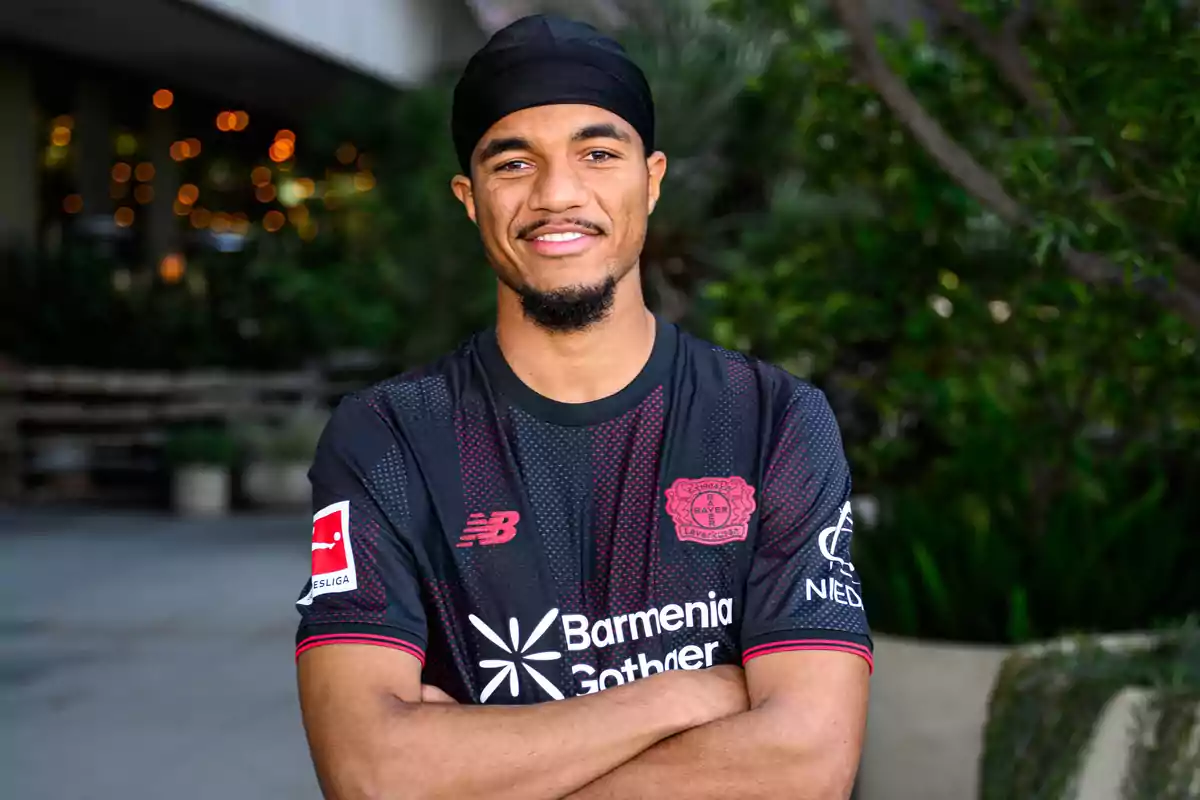 Jugador de fútbol con camiseta negra del Bayer Leverkusen posando con los brazos cruzados y sonriendo al aire libre Jugador de fútbol con camiseta negra del Bayer Leverkusen posando con los brazos cruzados y sonriendo al aire libre