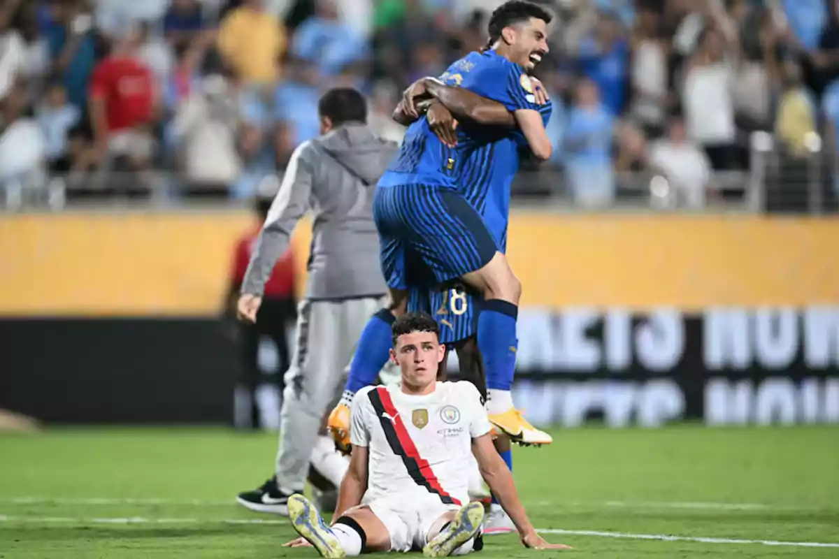 Un jugador con uniforme blanco está sentado en el césped con expresión de decepción mientras dos jugadores con uniforme azul celebran abrazados detrás de él en un estadio lleno de público. Un jugador con uniforme blanco está sentado en el césped con expresión de decepción mientras dos jugadores con uniforme azul celebran abrazados detrás de él en un estadio lleno de público.