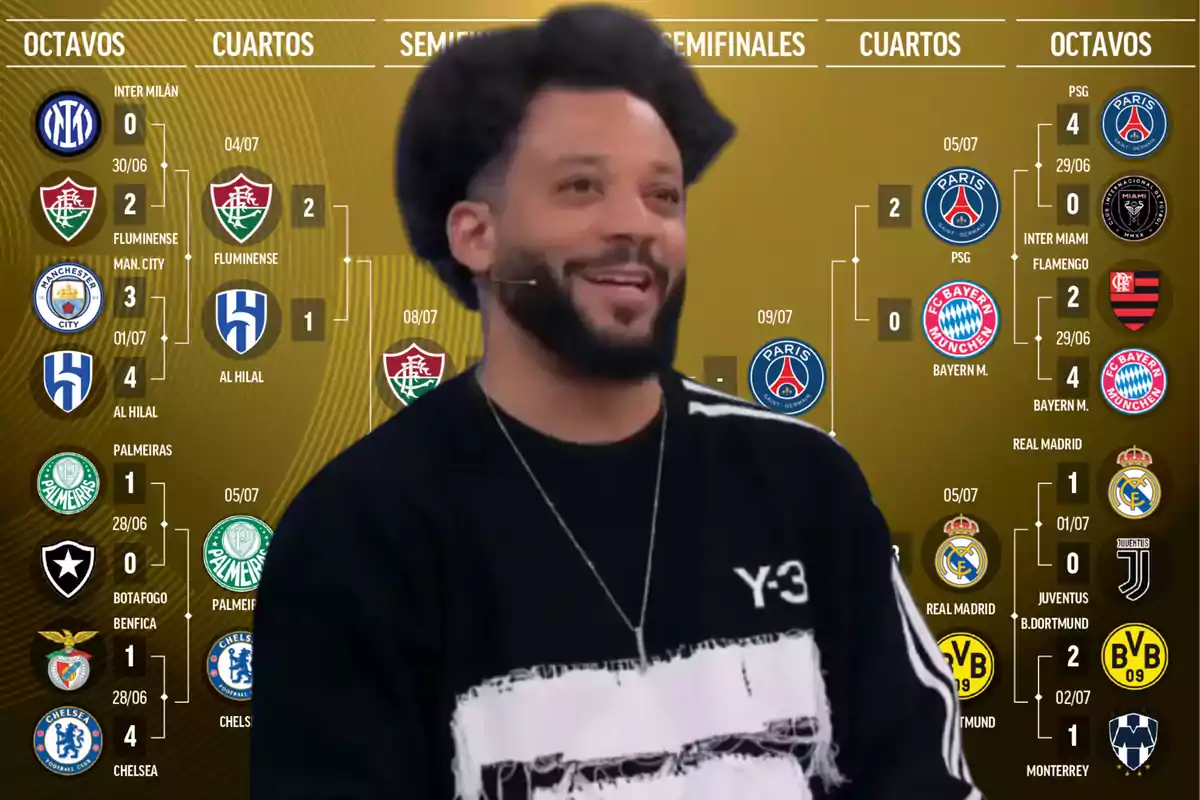 Un hombre sonriente con barba y cabello rizado lleva puesta una camiseta negra con detalles blancos, al fondo se observa un gráfico de un torneo de fútbol con los logotipos de varios equipos y los resultados de los partidos en diferentes fases del campeonato.