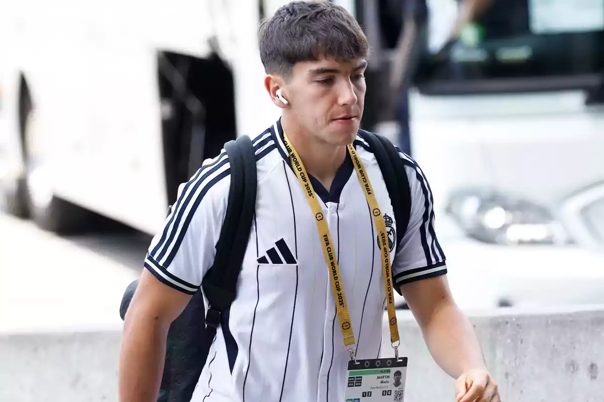 Joven con camiseta de fútbol blanca con rayas azules y mochila caminando junto a un autobús mientras lleva auriculares y una acreditación colgada al cuello Joven con camiseta de fútbol blanca con rayas azules y mochila caminando junto a un autobús mientras lleva auriculares y una acreditación colgada al cuello