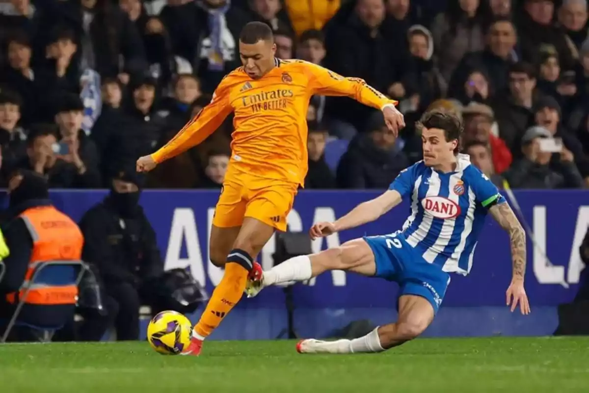Dos futbolistas disputan el balón durante un partido, uno con uniforme naranja y otro con uniforme azul y blanco, mientras el público observa desde las gradas. Dos futbolistas disputan el balón durante un partido, uno con uniforme naranja y otro con uniforme azul y blanco, mientras el público observa desde las gradas.