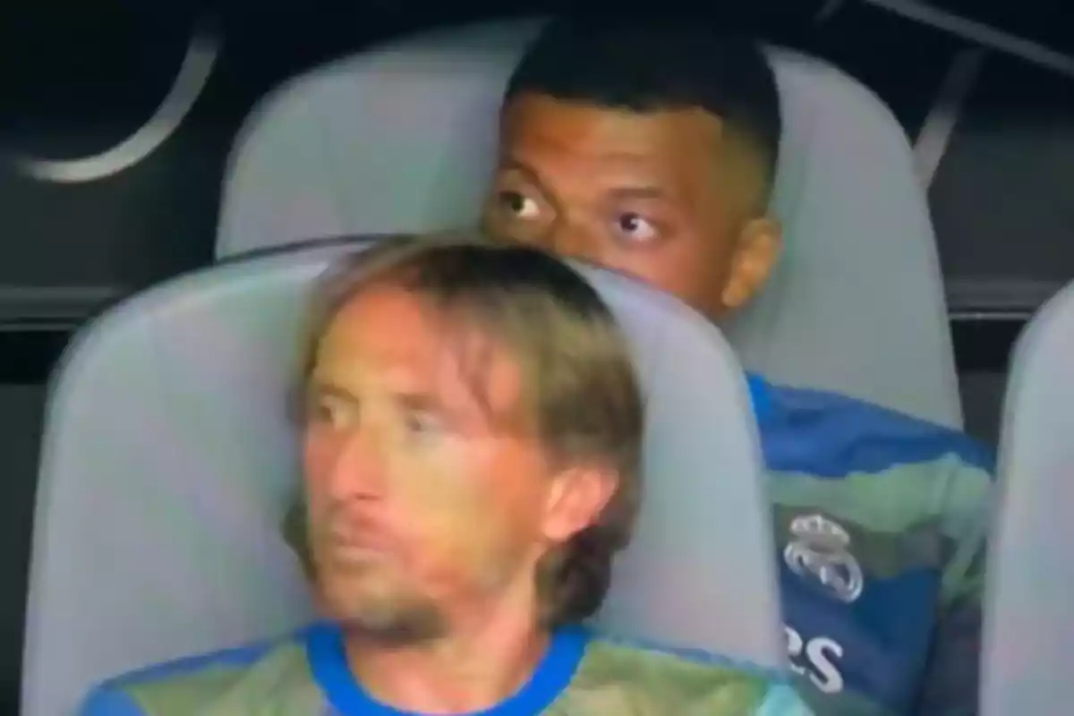 Dos jugadores del Real Madrid sentados en la banca, uno mirando hacia un lado y el otro asomándose por detrás del asiento.