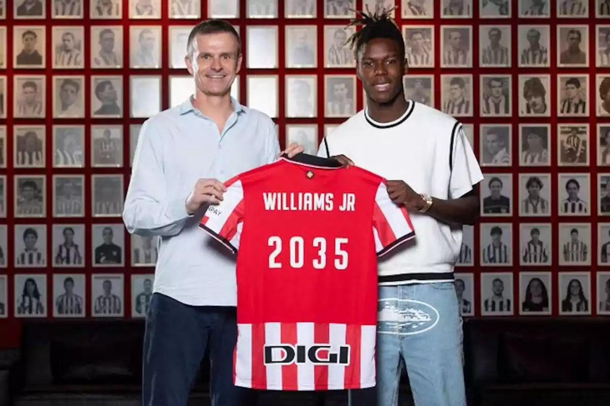 Two people pose smiling, holding a red and white soccer jersey with the name Williams Jr and the number 2035, in front of a wall filled with black-and-white framed portraits. Two people pose smiling, holding a red and white soccer jersey with the name Williams Jr and the number 2035, in front of a wall filled with black-and-white framed portraits.