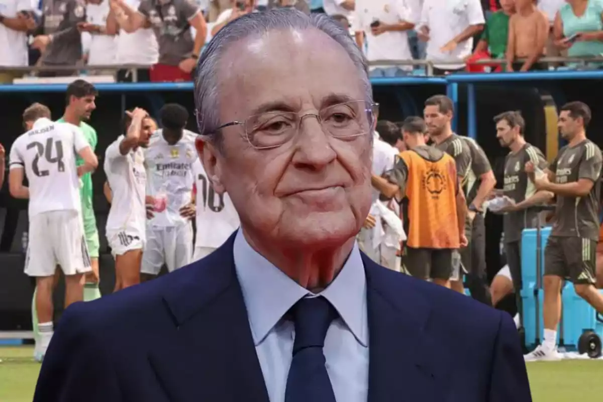 Gray-haired man with glasses in a dark suit standing in front of a group of players and coaching staff in a stadium