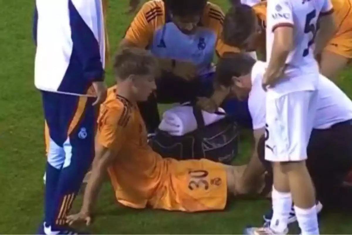 Un jugador de fútbol con uniforme naranja está sentado en el césped mientras recibe atención médica de varias personas durante un partido Un jugador de fútbol con uniforme naranja está sentado en el césped mientras recibe atención médica de varias personas durante un partido