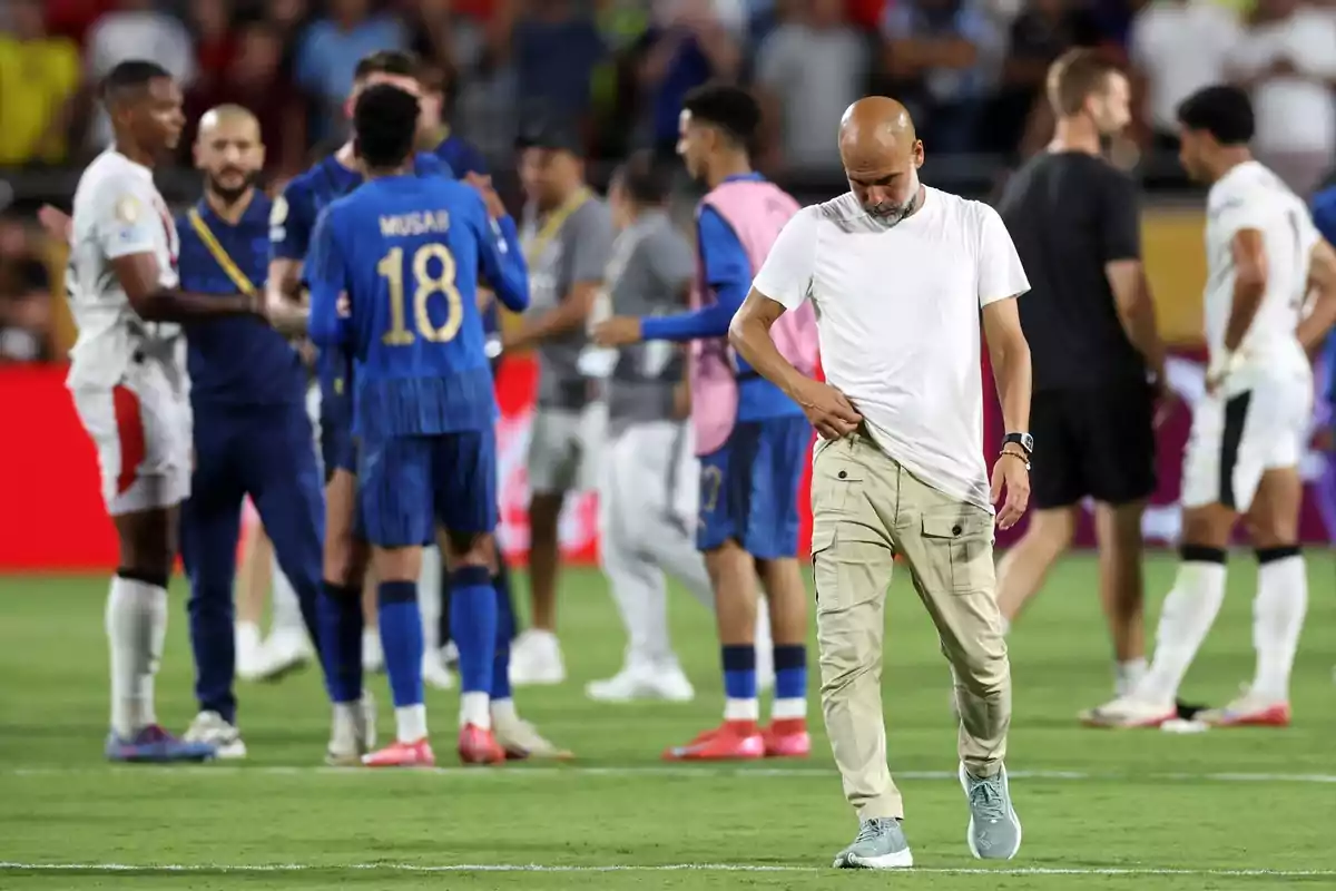 Un entrenador de fútbol con expresión seria camina cabizbajo sobre el campo mientras jugadores de ambos equipos se saludan al fondo tras un partido Un entrenador de fútbol con expresión seria camina cabizbajo sobre el campo mientras jugadores de ambos equipos se saludan al fondo tras un partido