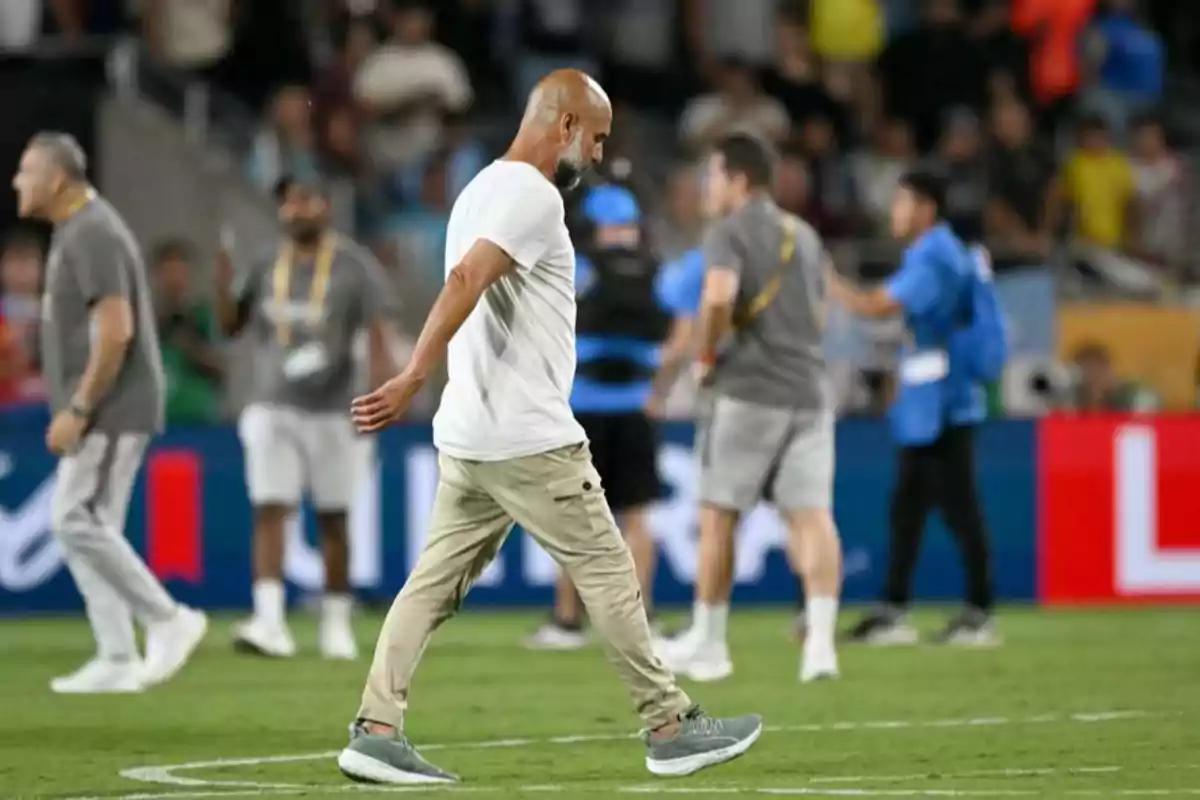 Hombre calvo con barba y camiseta blanca camina cabizbajo sobre el césped de un estadio mientras otras personas se ven desenfocadas al fondo
