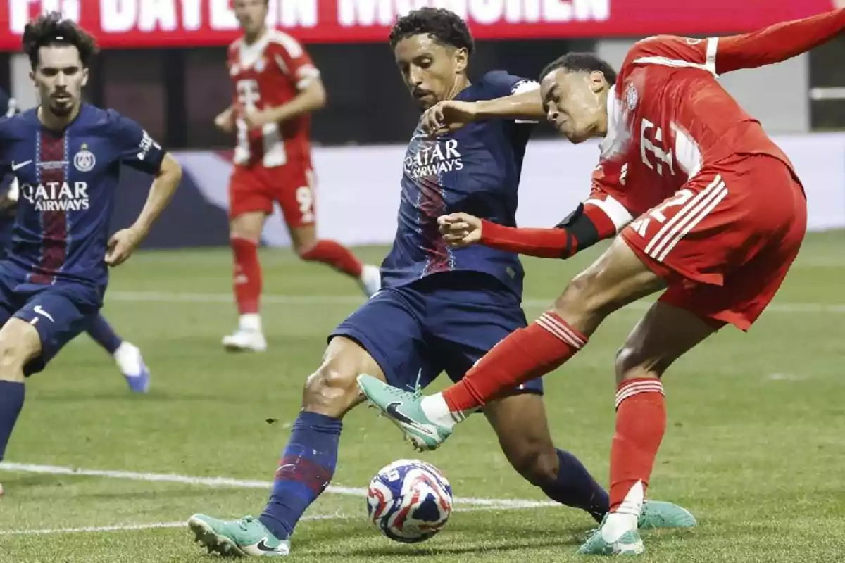 Two players compete for the ball during a match between teams in blue and red uniforms, while other players watch the play in the background. Two players compete for the ball during a match between teams in blue and red uniforms, while other players watch the play in the background.