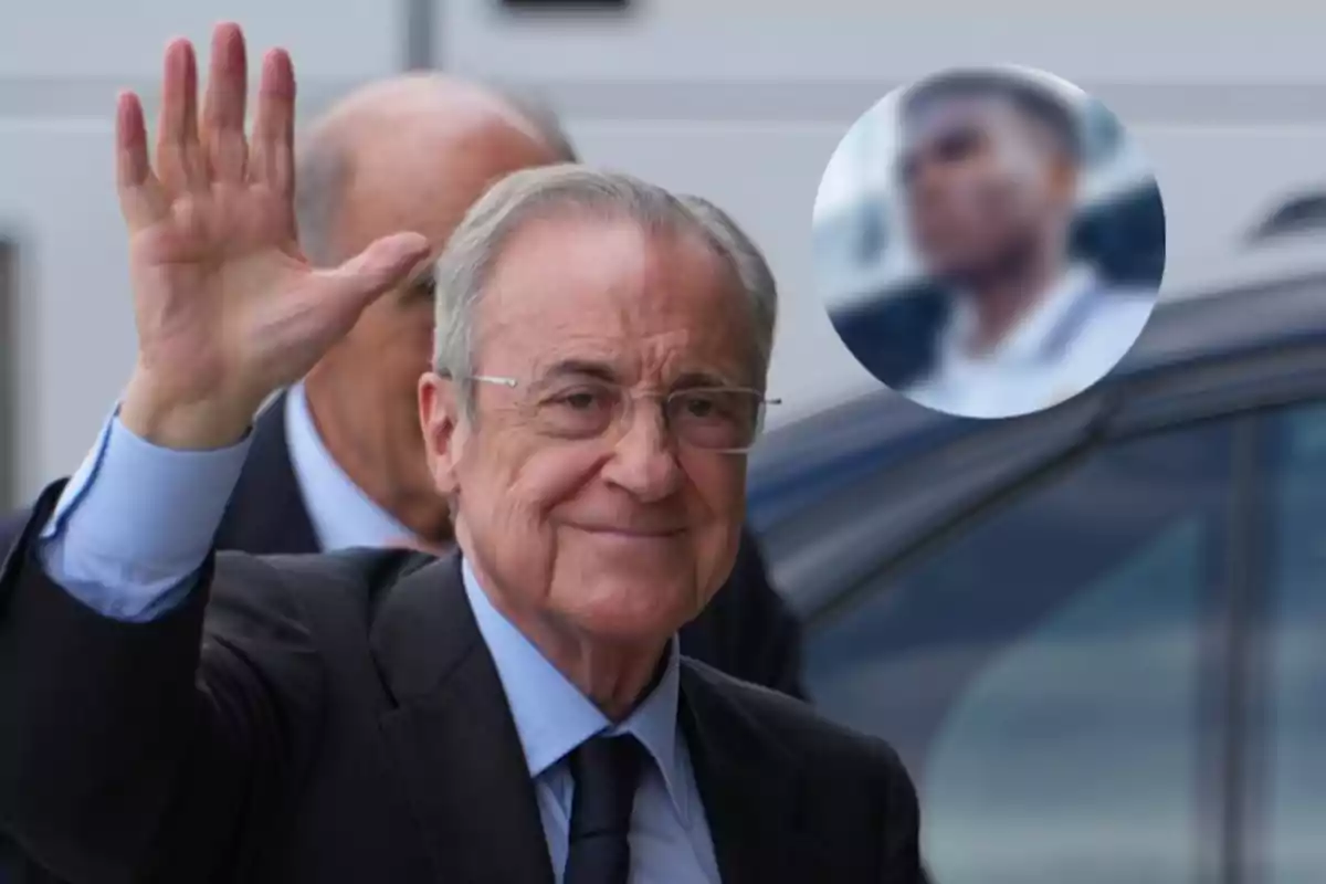 Older man with glasses and a dark suit waves while smiling; in the background, there is a circle with the blurred image of another person.