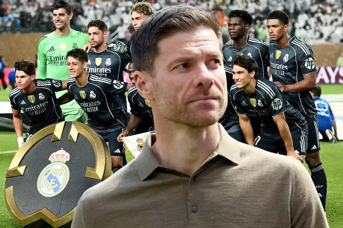 Un hombre en primer plano con expresión seria y al fondo un equipo de fútbol vestido con uniforme negro posando para una foto en el campo junto a un escudo grande del Real Madrid