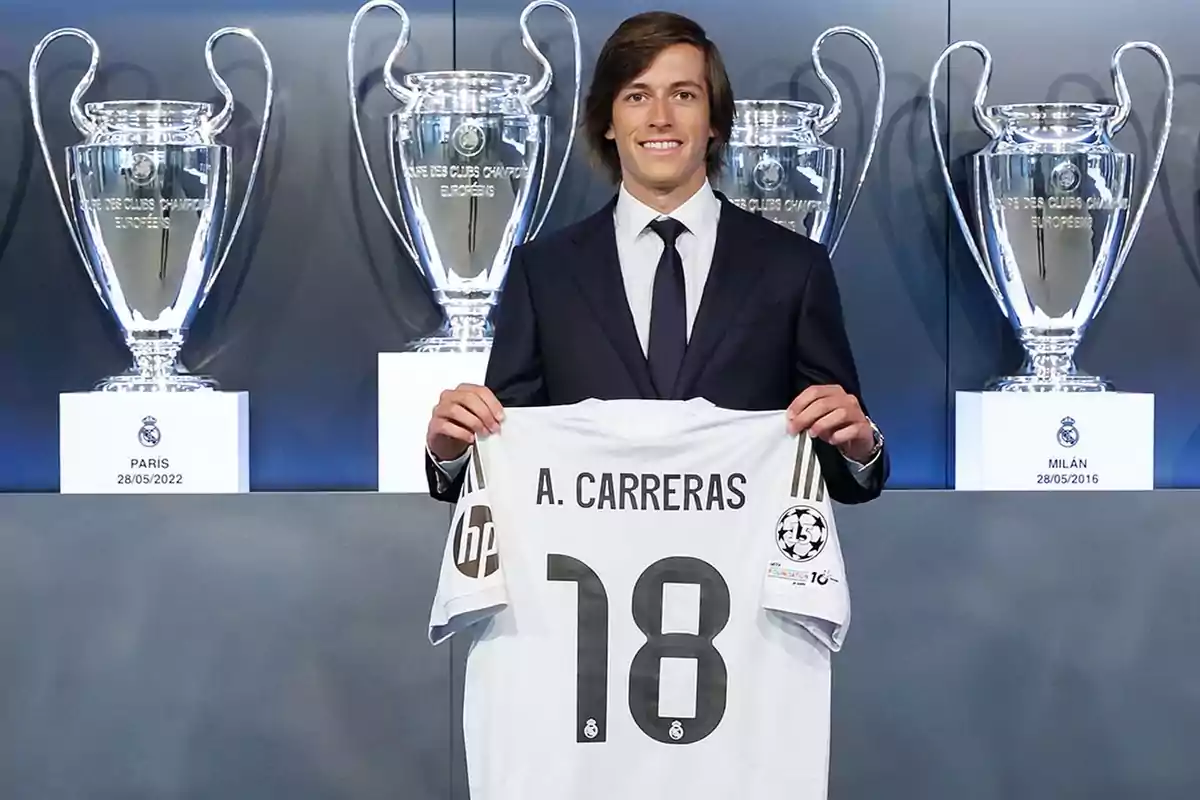 Un hombre con traje sostiene una camiseta de fútbol con el nombre A. Carreras y el número 18 frente a varios trofeos grandes de la Champions League. Un hombre con traje sostiene una camiseta de fútbol con el nombre A. Carreras y el número 18 frente a varios trofeos grandes de la Champions League.