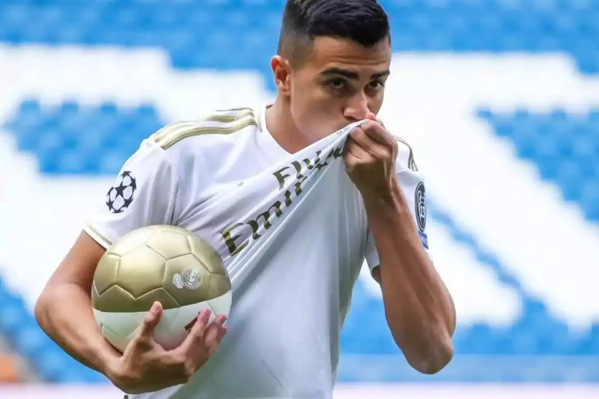 Futbolista con uniforme blanco besando el escudo de su camiseta y sosteniendo un balón dorado en un estadio Futbolista con uniforme blanco besando el escudo de su camiseta y sosteniendo un balón dorado en un estadio