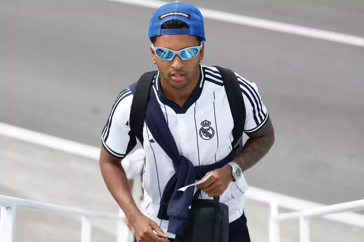 Player with sunglasses and a blue cap worn backwards going up some stairs, wearing a Real Madrid jersey and a black backpack Player with sunglasses and a blue cap worn backwards going up some stairs, wearing a Real Madrid jersey and a black backpack