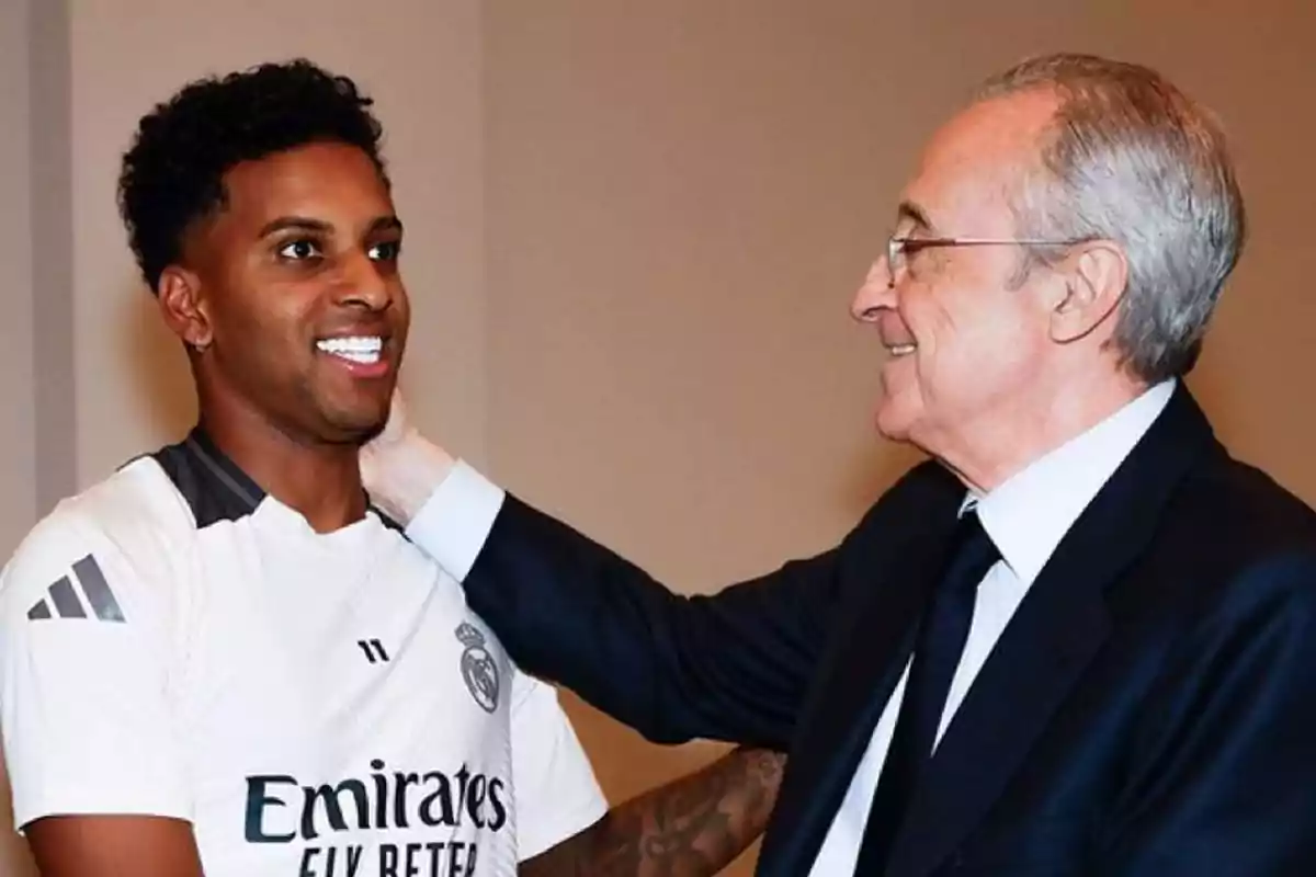 Un joven con camiseta de fútbol blanca sonríe mientras un hombre mayor de traje lo saluda amablemente colocando una mano en su cuello Un joven con camiseta de fútbol blanca sonríe mientras un hombre mayor de traje lo saluda amablemente colocando una mano en su cuello