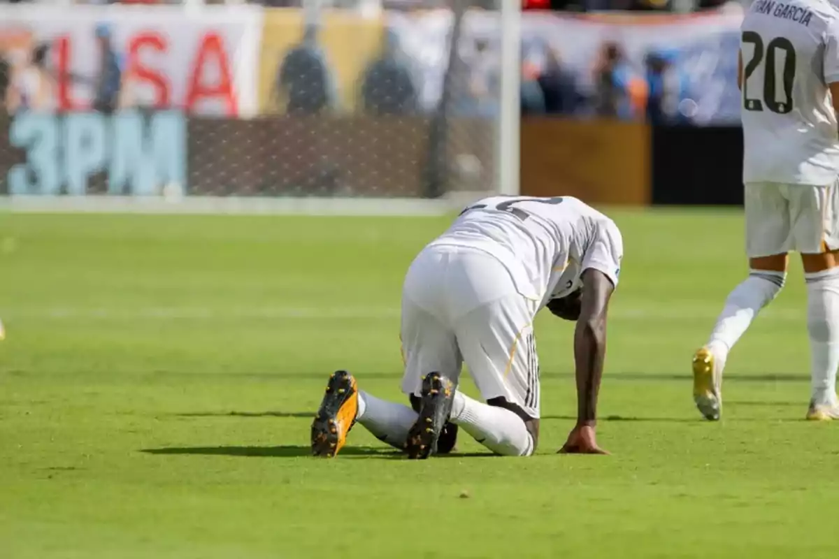 Futbolista arrodillado en el césped durante un partido mientras otro jugador camina cerca Futbolista arrodillado en el césped durante un partido mientras otro jugador camina cerca