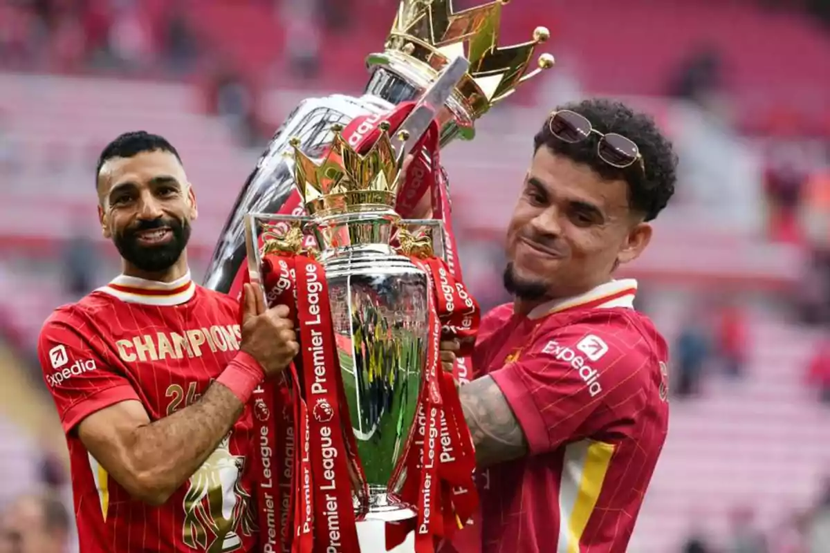Dos futbolistas del Liverpool sonríen y levantan el trofeo de la Premier League en el estadio, vistiendo camisetas rojas de campeones.