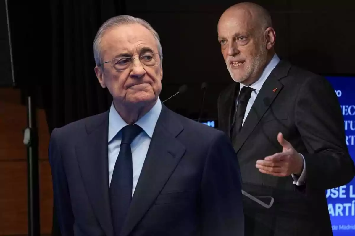 Two men in suits at a formal event, one in front with a serious expression and another in the background speaking into a microphone.