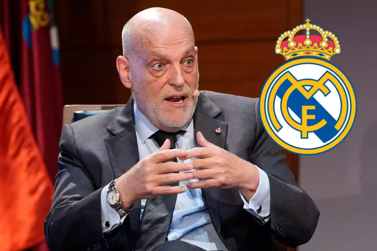 Bald man in a suit gesturing while speaking with the Real Madrid crest next to him