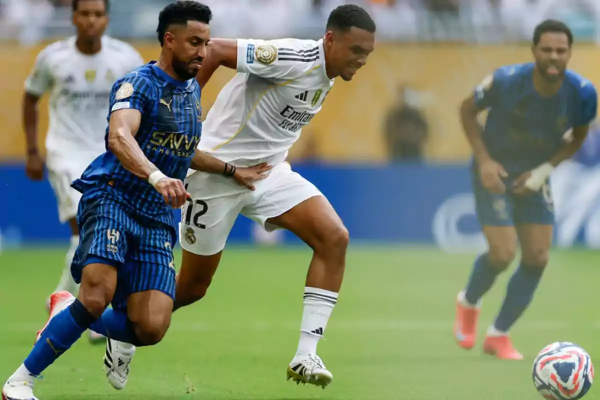 Dos futbolistas disputan el balón durante un partido mientras otros jugadores observan la jugada en el fondo Dos futbolistas disputan el balón durante un partido mientras otros jugadores observan la jugada en el fondo