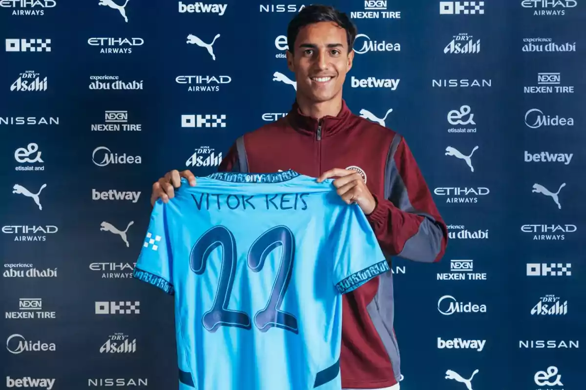 A smiling player holds a blue jersey with the name Vitor Reis and the number 22 in front of a background with sponsor logos. A smiling player holds a blue jersey with the name Vitor Reis and the number 22 in front of a background with sponsor logos.